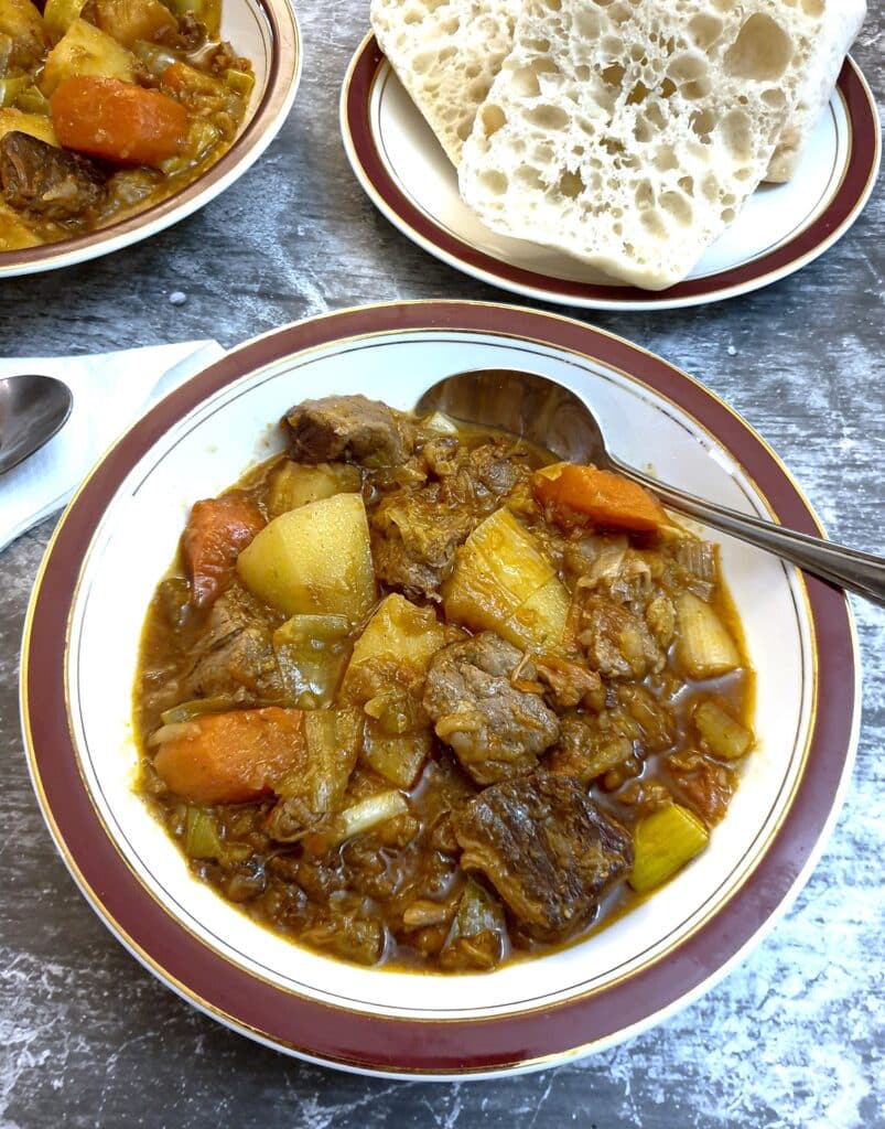 A bowl of lamb stew in a red-rimmed plate in front of a plate of sour dough bread.