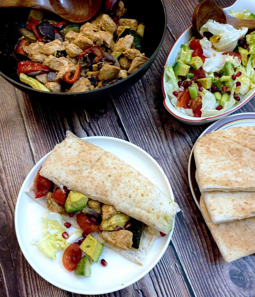 Overhead shot of a pita bread filled with Moroccan chicken on a white plate, next to a bowl of salad and a pan of chicken.