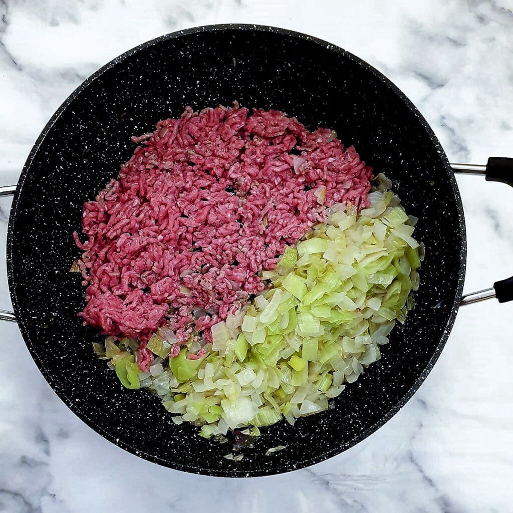 Ground beef being softened with onions and leeks in a frying pan.