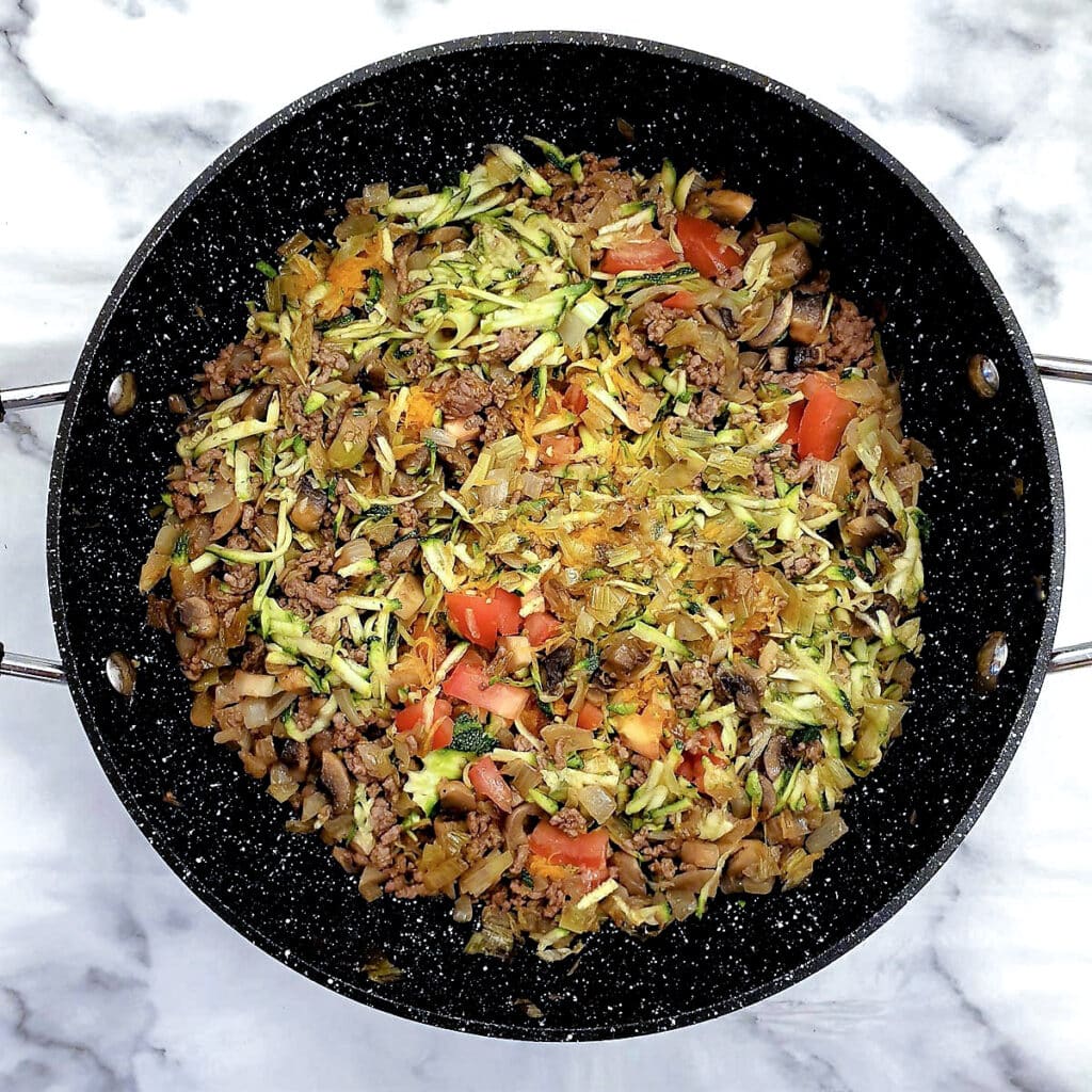 Grated courgettes, carrots and tomatoes frying with mince and onions in a frying pan.