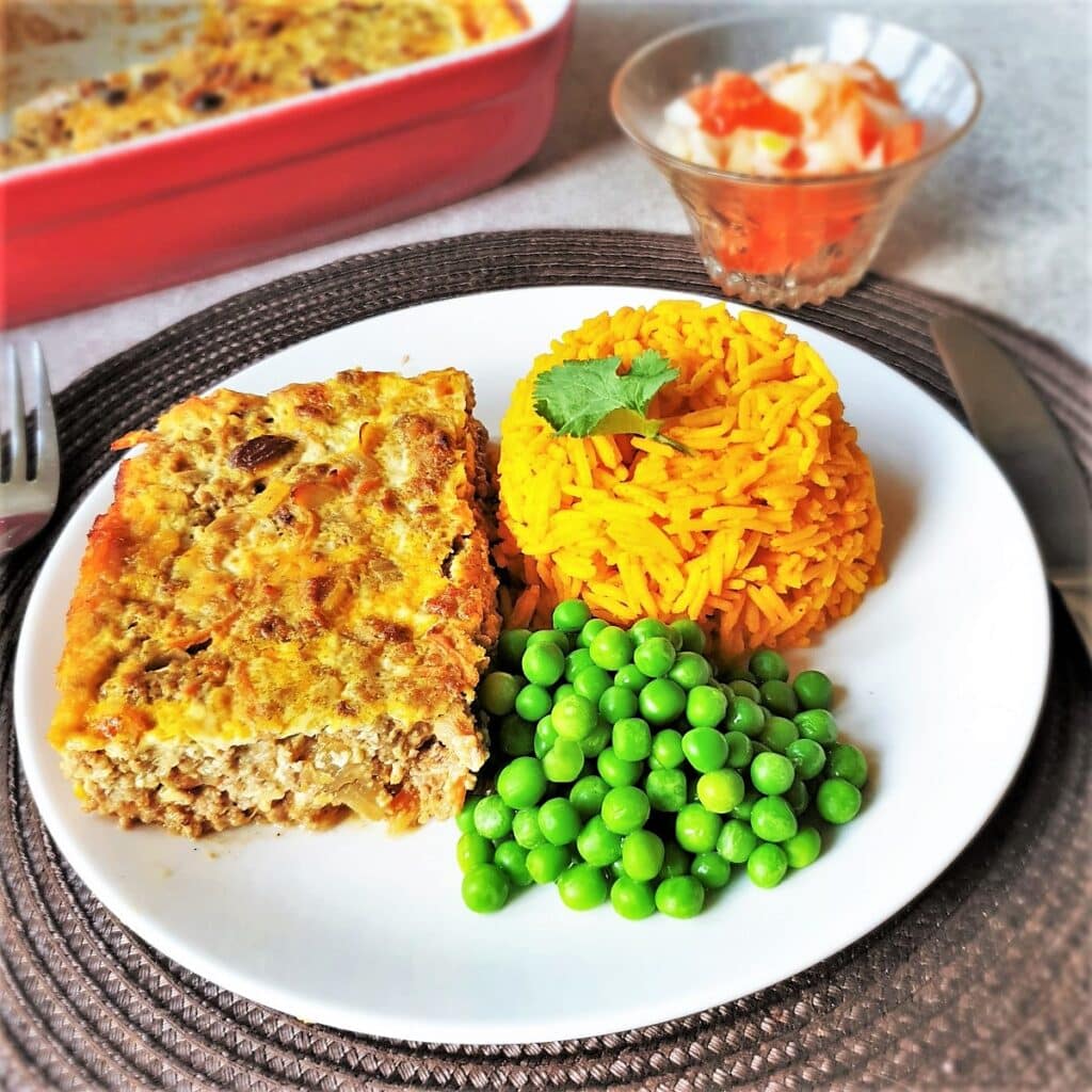 A helping of bobotie on a white place alongside yellow rice and peas.