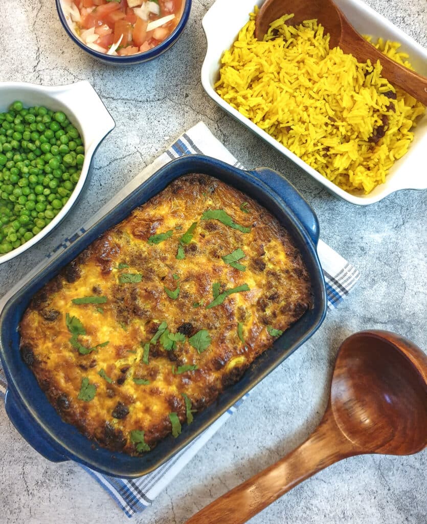 A blue serving dish of bobotie next to a bowl of yellow rice and a bowl of peas.