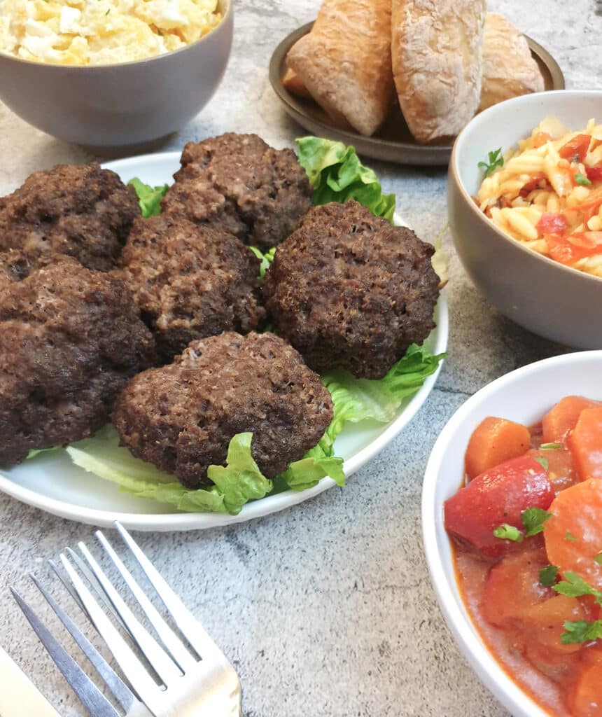 6 homemade boerewors patties on a table alongside bowls of salad and bread rolls.