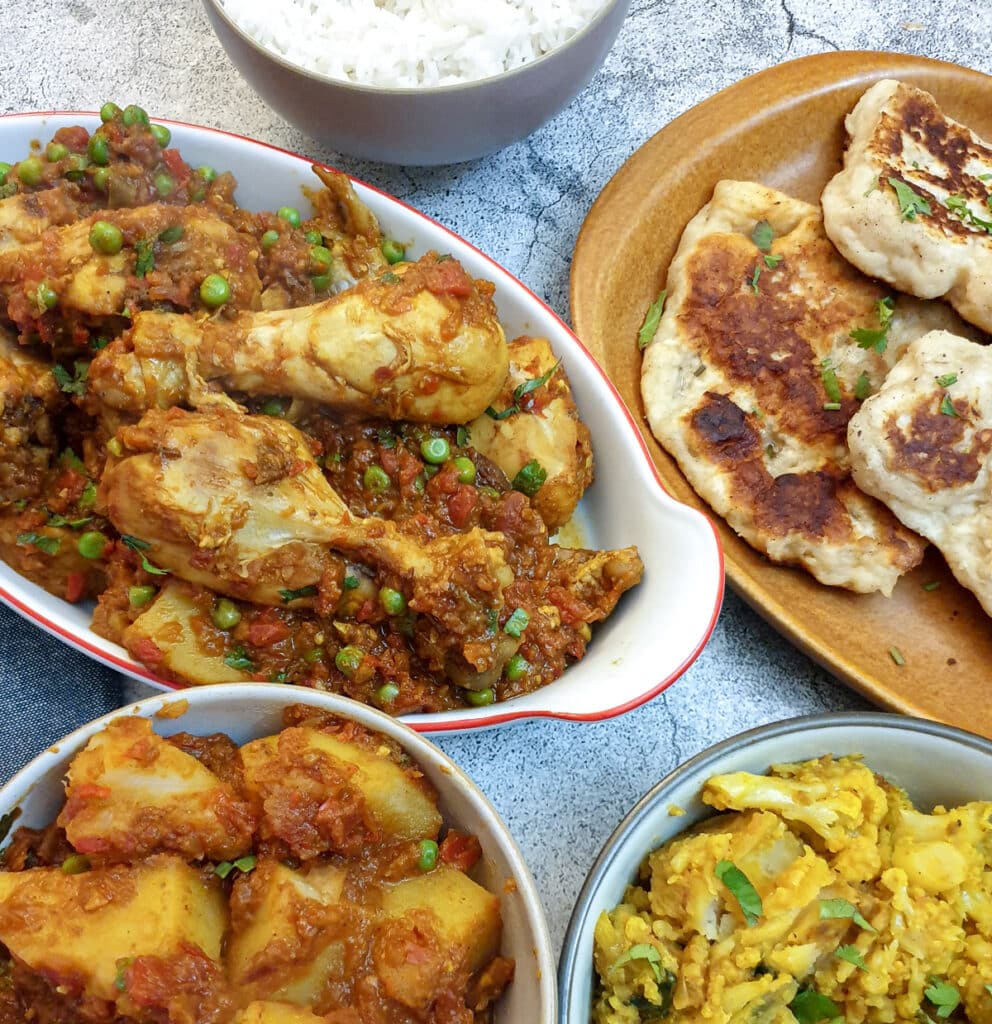 A dish of chicken bhuna on a table with bombay potatoes, naan bread rice and aloo gobi.