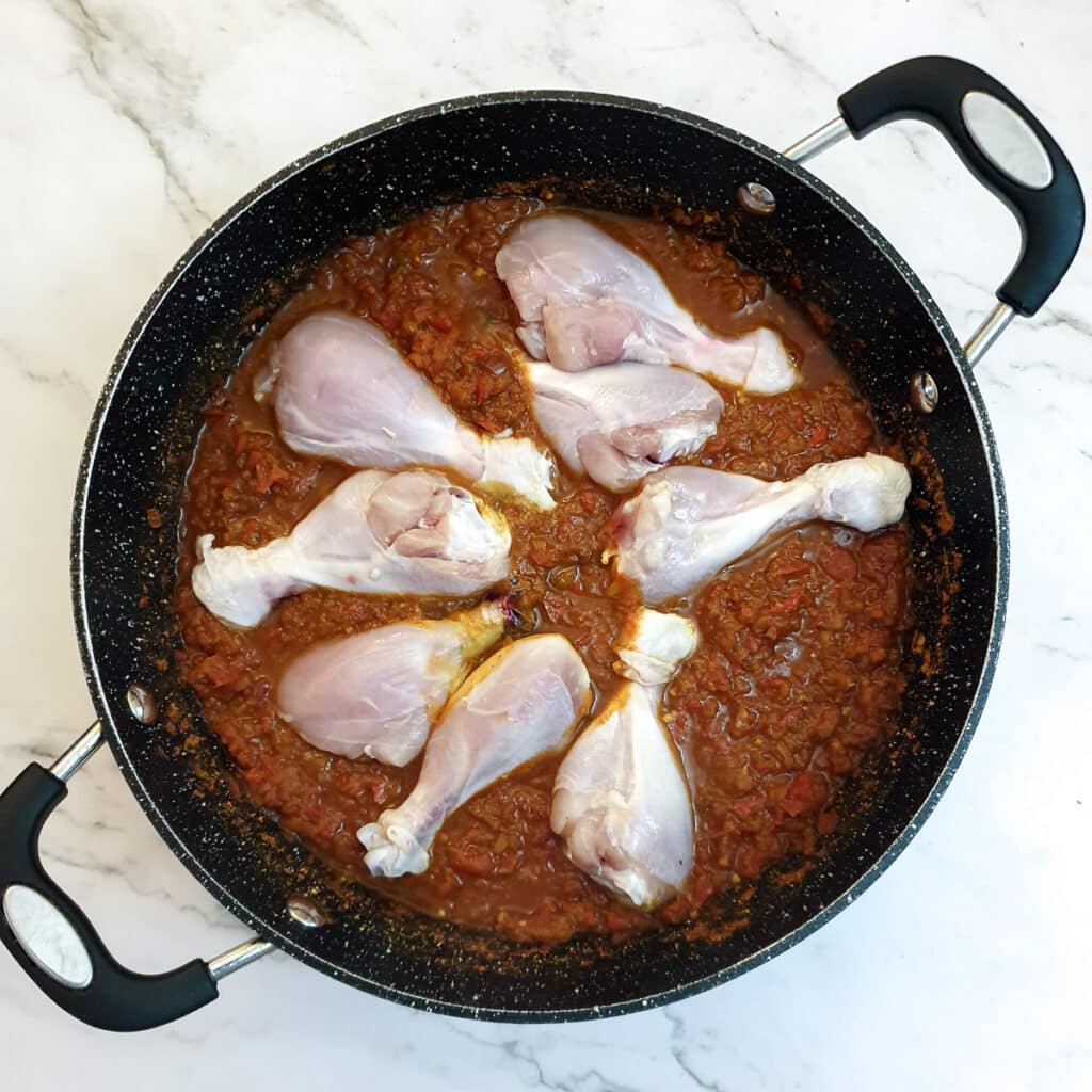 Chicken drumsticks in a frying pan with red pepper and onion curry paste.