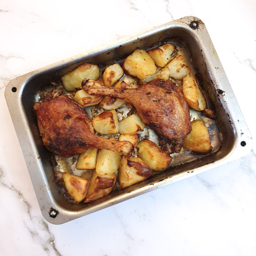Two crispy duck legs with crispy roast potatoes in a roasting pan.