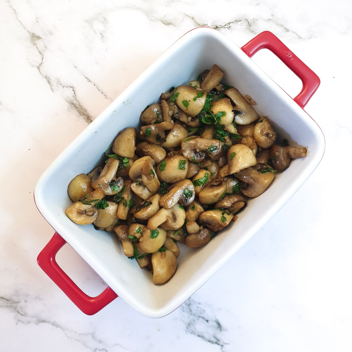 A white serving dish with red handles filled with garlic mushrooms.