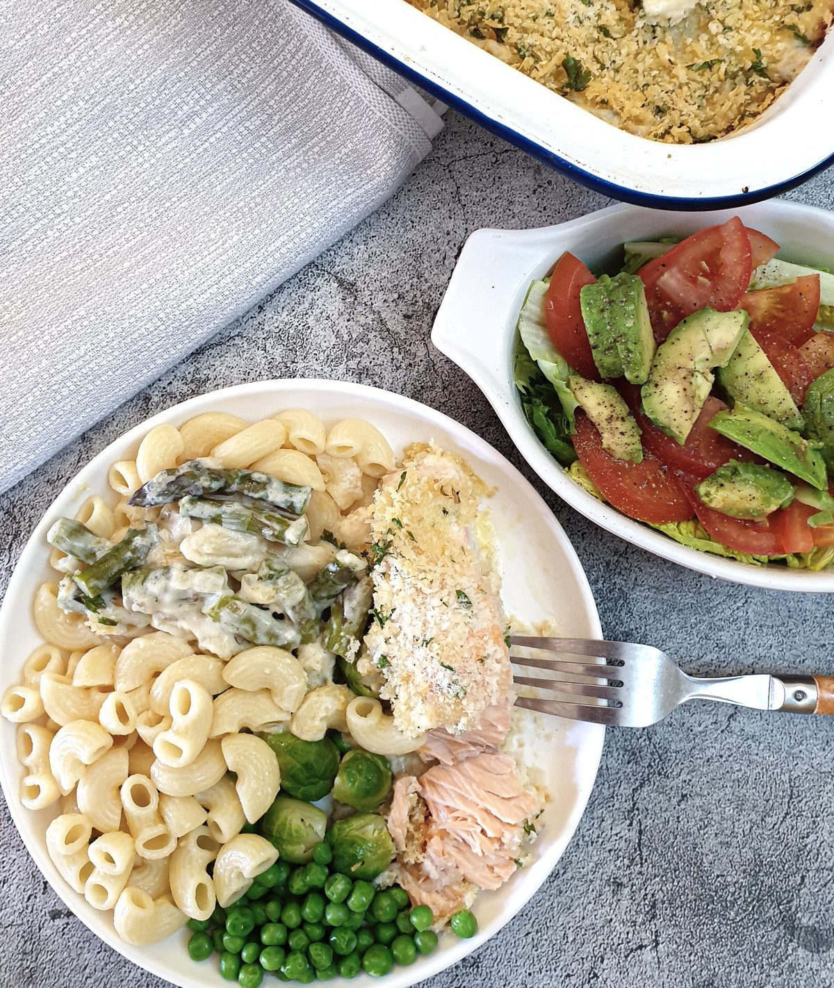 A slice of salmon topped with breadcrumbs on a white plate with buttered noodles and vegetables.