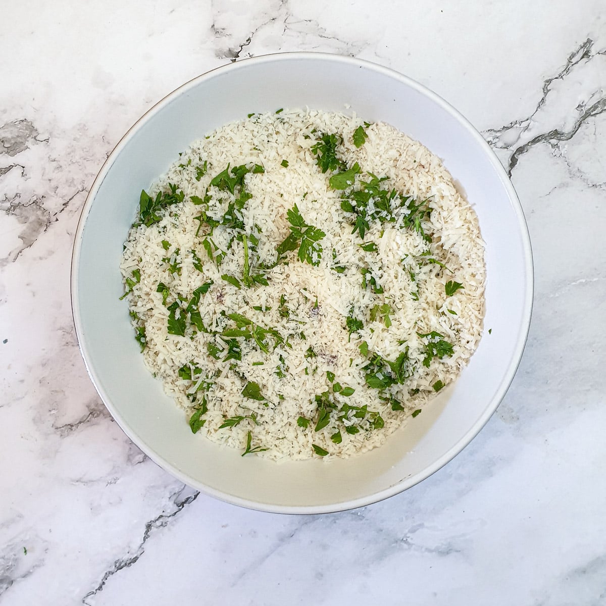 Grated parmesan cheese, breadcrumbs and chopped parsley combined in a white bowl.