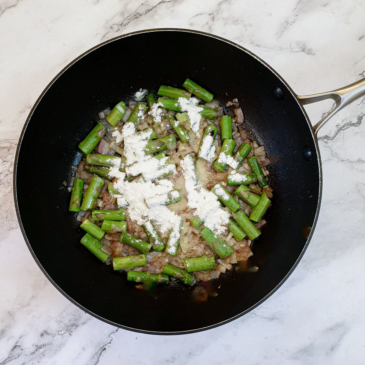 Flour sprinkled over shallots and asparagus stalks in a frying pan.