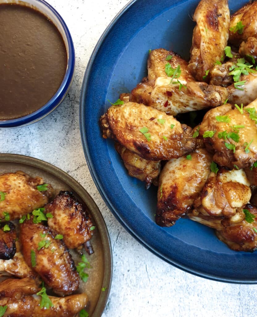 A blue serving dish of sticky chicken wings next to a plate of wings.