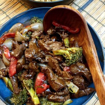 A blue dish filled with Szechuan beef stir fry with a wooden serving spoon.