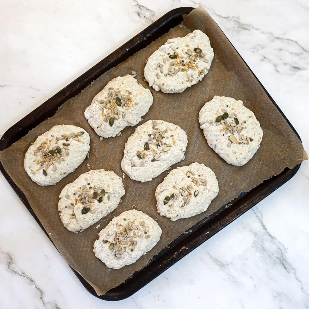 8 unbaked seeded breadrolls on a baking tray.