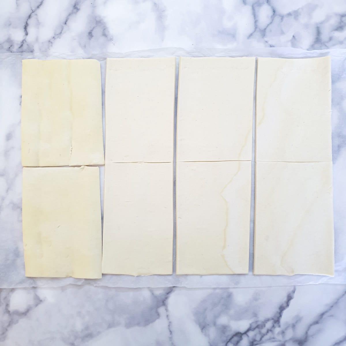 An unbaked roll of puff pastry unrolled on a table and cut into 8 rectangles.