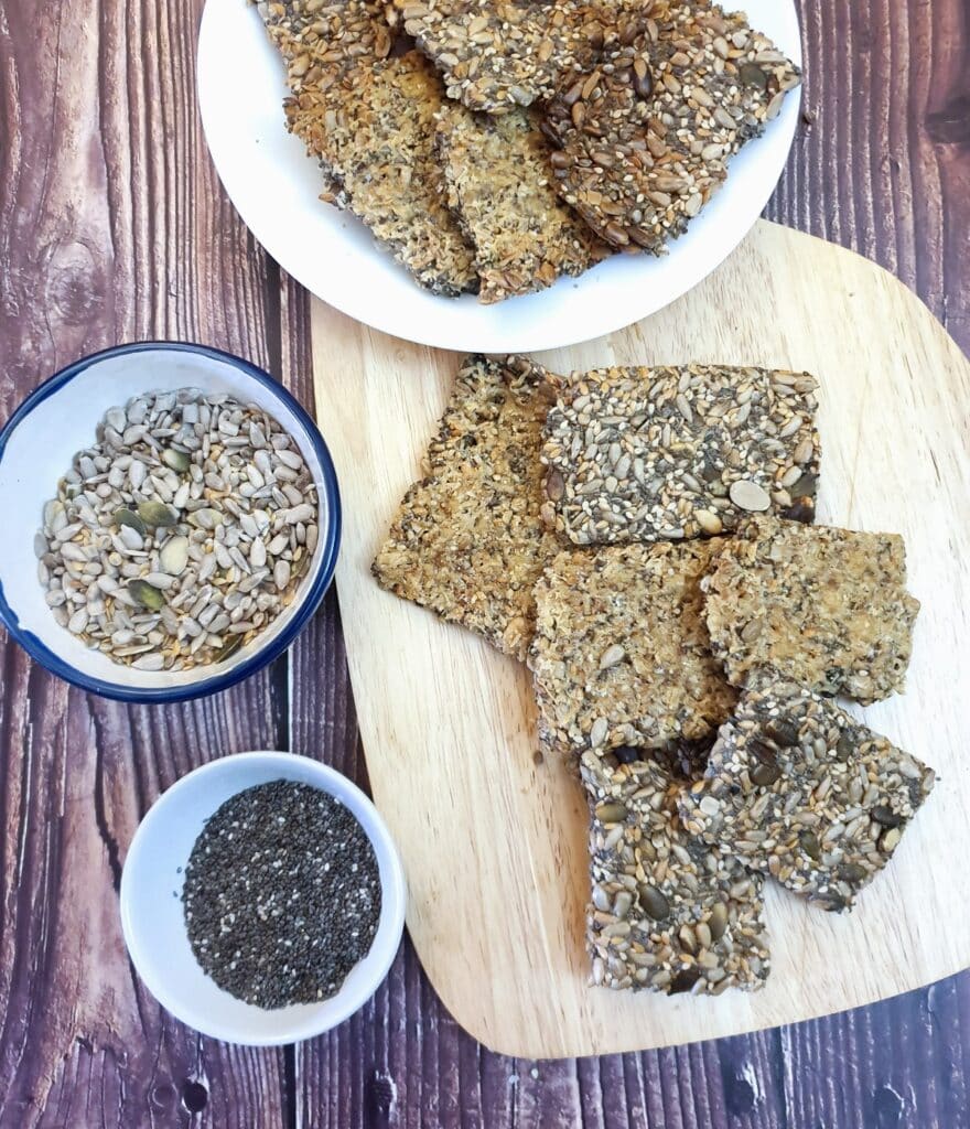 A wooden board with a pile of crispy seeded crackers next to two bowls of seeds.