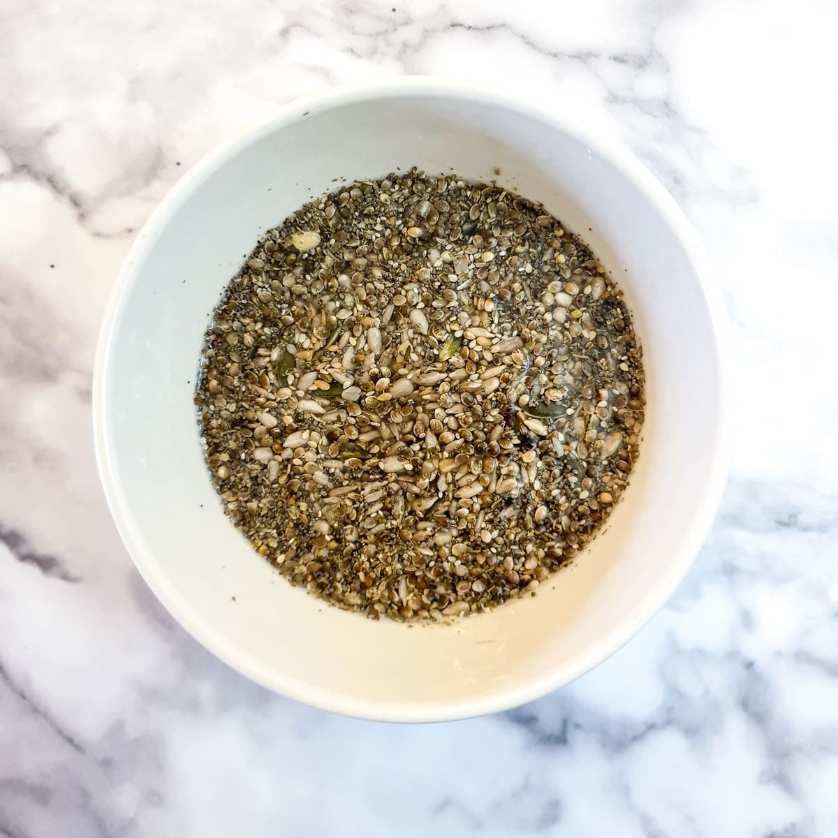 A white bowl containing mixed seeds soaking in a cup of water.