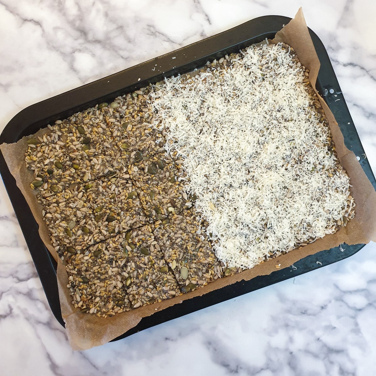 A baking tray of seeded cracker mixture, half of which has been covered in grated parmesan cheese.