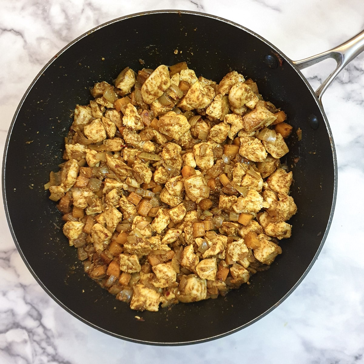 Spices and chicken pieces mixed with onions and carrots in a frying pan.