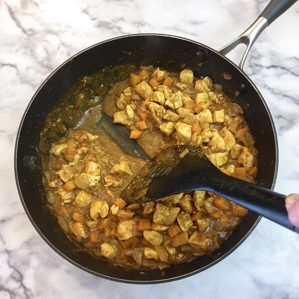 A spatula being drawn through a pan of curried chicken showing the consistency of the sauce.