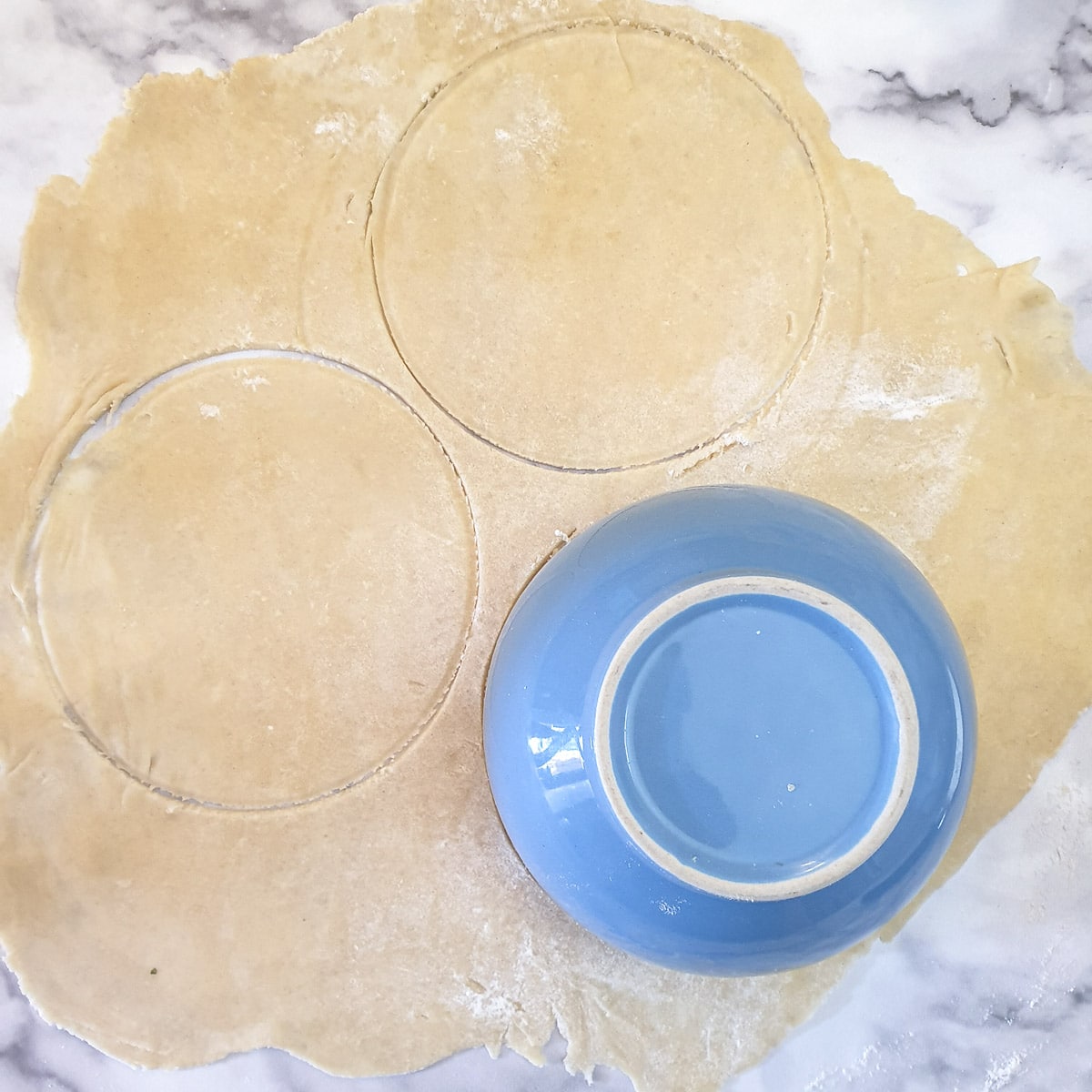 A piece of rolled-out pastry being cut into circles with the aid of a blue bowl.