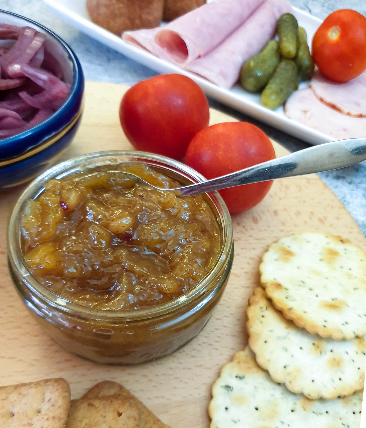 A dish of mango chutney on a wooden board, surrounded by cold meat slices, tomatoes and crackers.