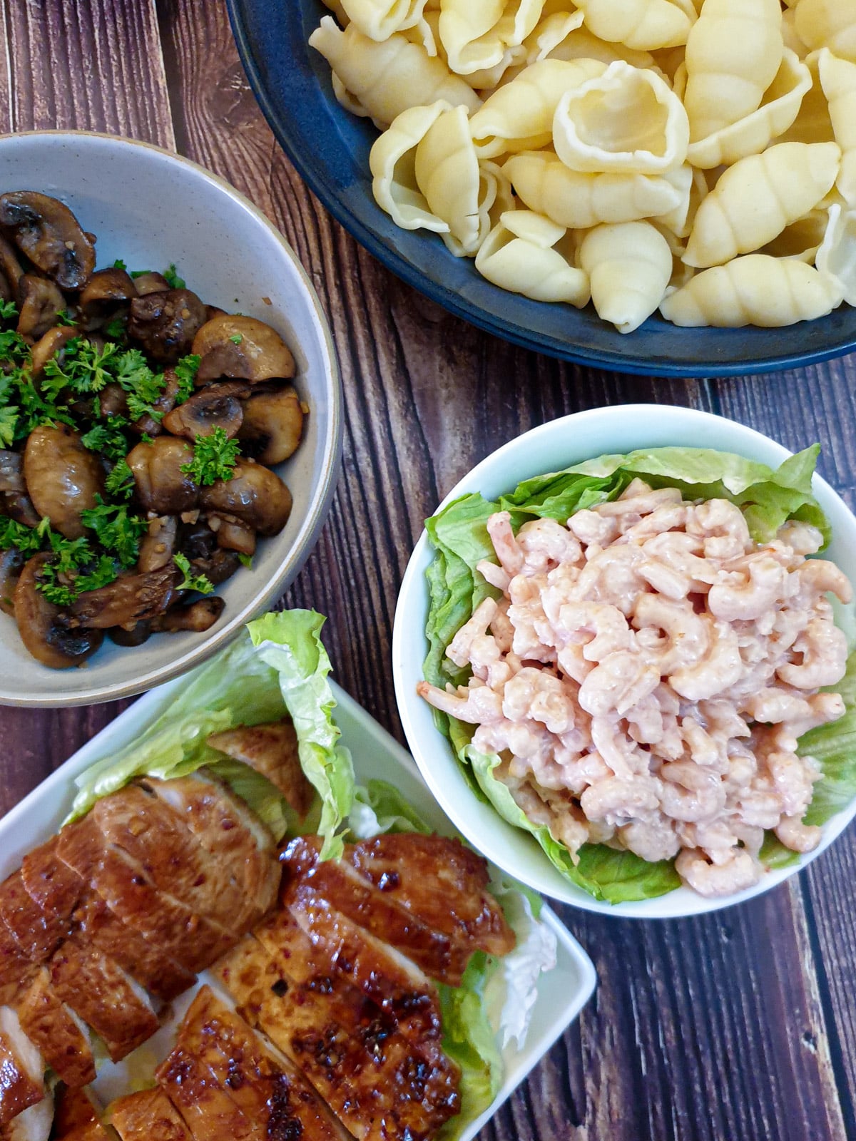 Honey garlic chicken breasts, garlic mushrooms, poorman's crayfish and a bowl of pasta on a wooden tabletop.