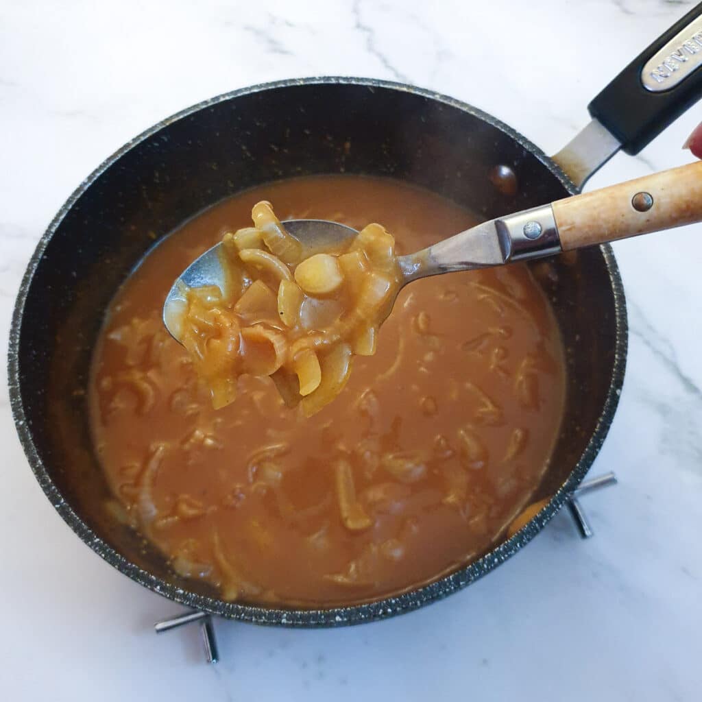 A spoonful of onion gravy held over a pan, showing the onions.