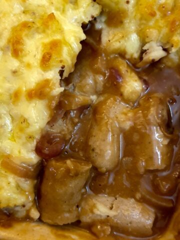 Pieces of sausages with onion gravy in the corner of a casserole dish, surrounded with cheesy mashed potatoes.