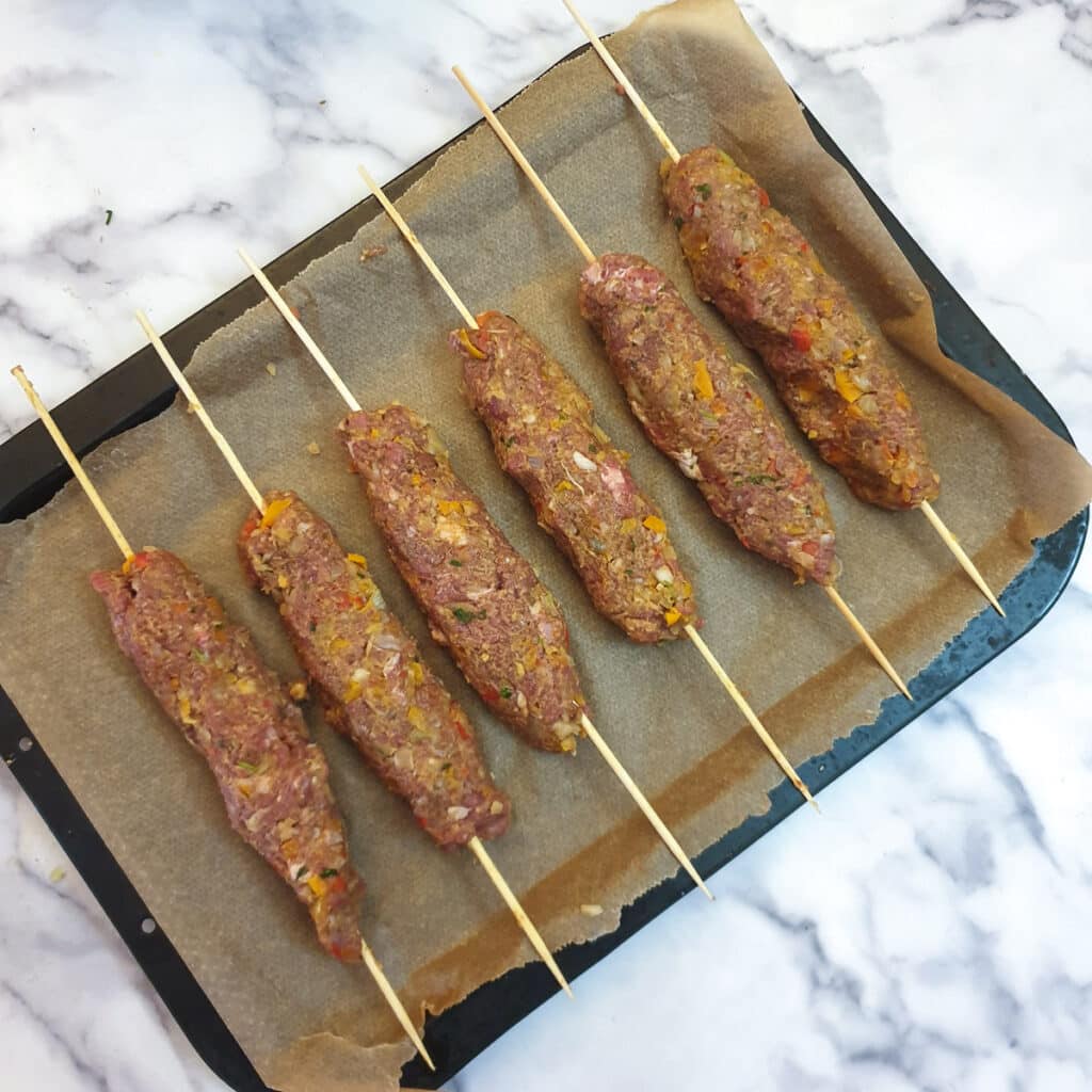 6 raw lam kofta on skewers arranged on a baking tray.