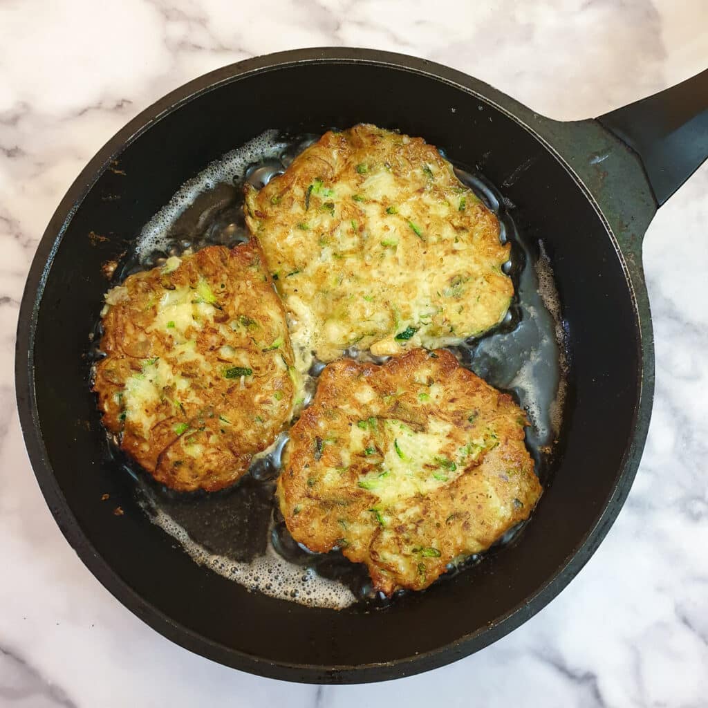 Three cheesy courgette fritters browning in a frying pan.