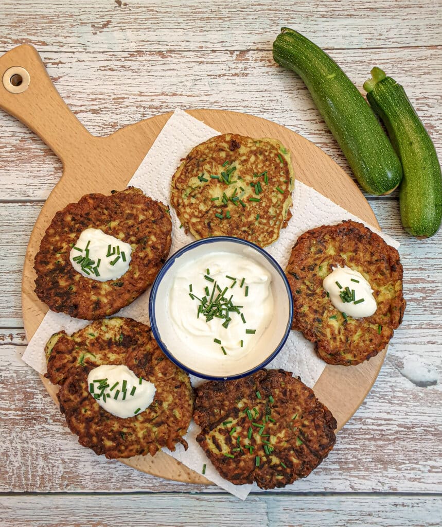 Overhead shot of 5 cheesy courgette fritters on a wooden board with a bowl of sour cream and chives in the centre.