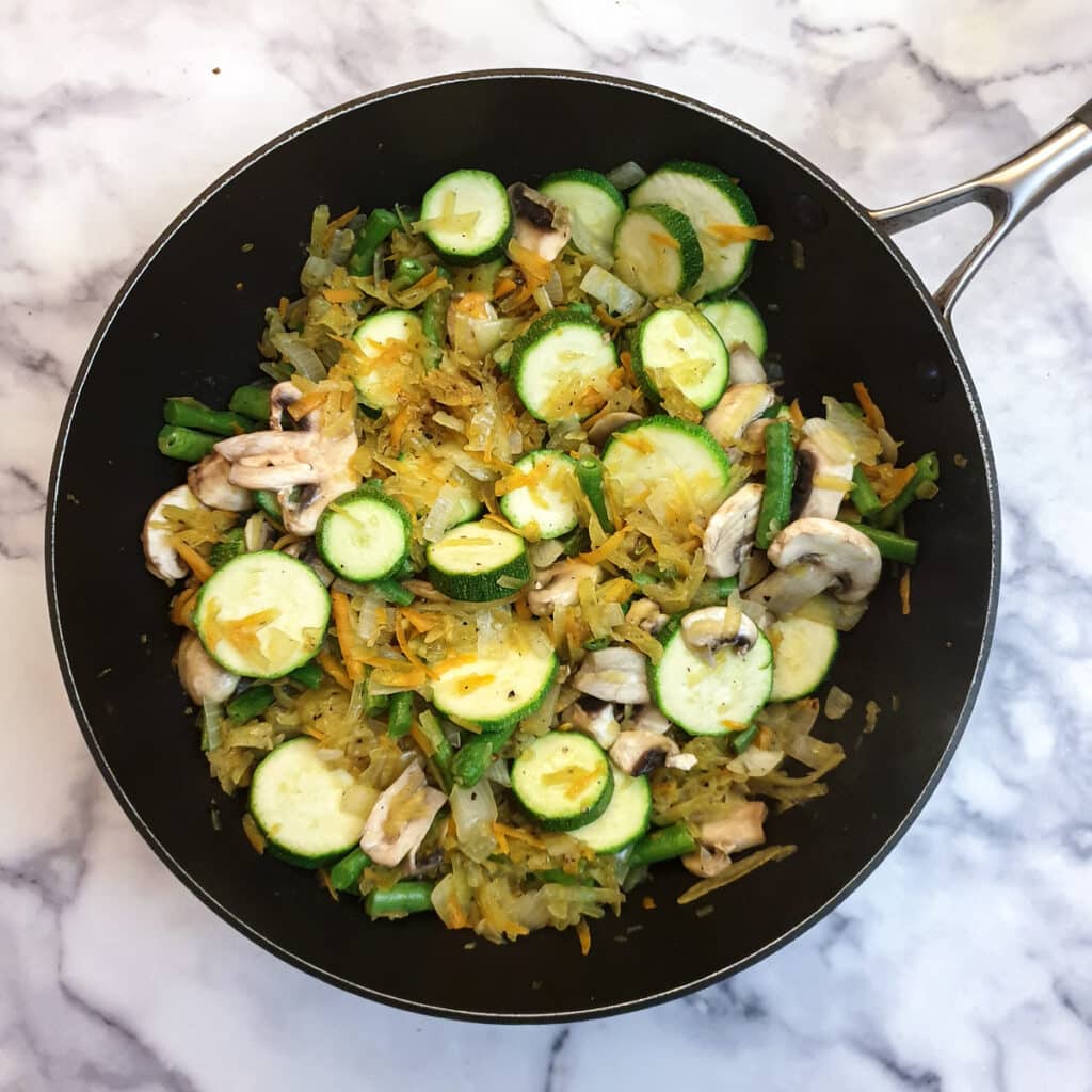 Mushrooms and courgettes added to the other vegetables in a frying pan.