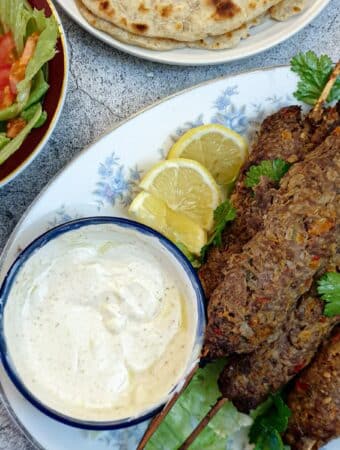 A pile of lamb kofte on a white plate with a dish of yoghurt sauce.