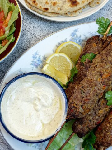 A pile of lamb kofte on a white plate with a dish of yoghurt sauce.