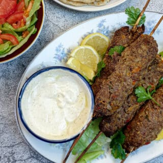 A pile of lamb kofte on a white plate with a dish of yoghurt sauce.