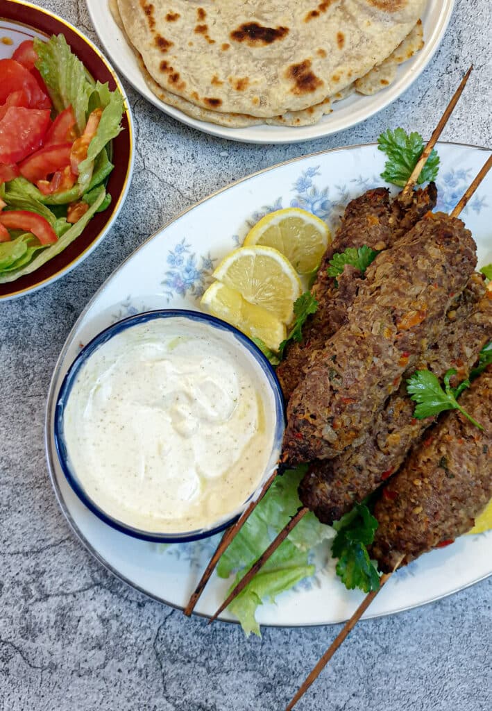 A pile of lamb kofte on a white plate with a dish of yoghurt sauce.