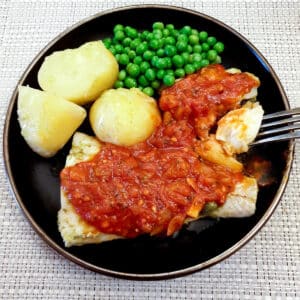 Oven-baked Mediterranean cod on a plate, covered with marinara sauce, with peas and boiled potatoes.