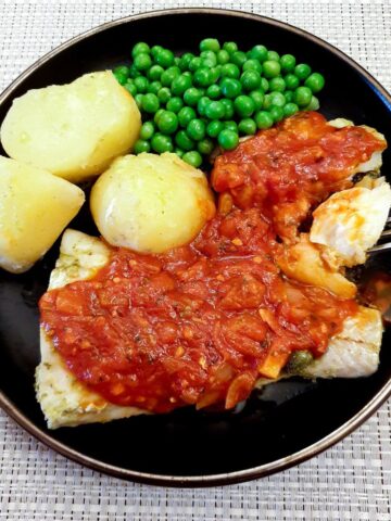 Oven-baked Mediterranean cod on a plate, covered with marinara sauce, with peas and boiled potatoes.