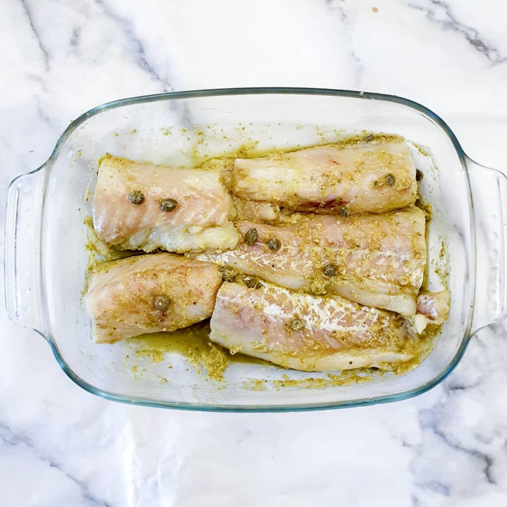 Cod fillets marinating in olive oil, capers and pesto in a glass dish.