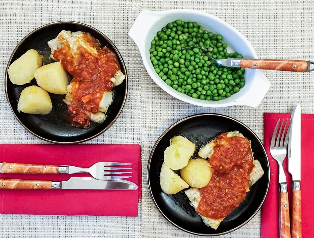 Two plates of mediterranean cod on a plate with potatoes, covered with marinara sauce alongside a dish of peas.