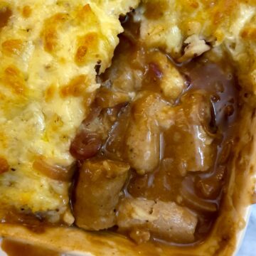 Pieces of sausages with onion gravy in the corner of a casserole dish, surrounded with cheesy mashed potatoes.