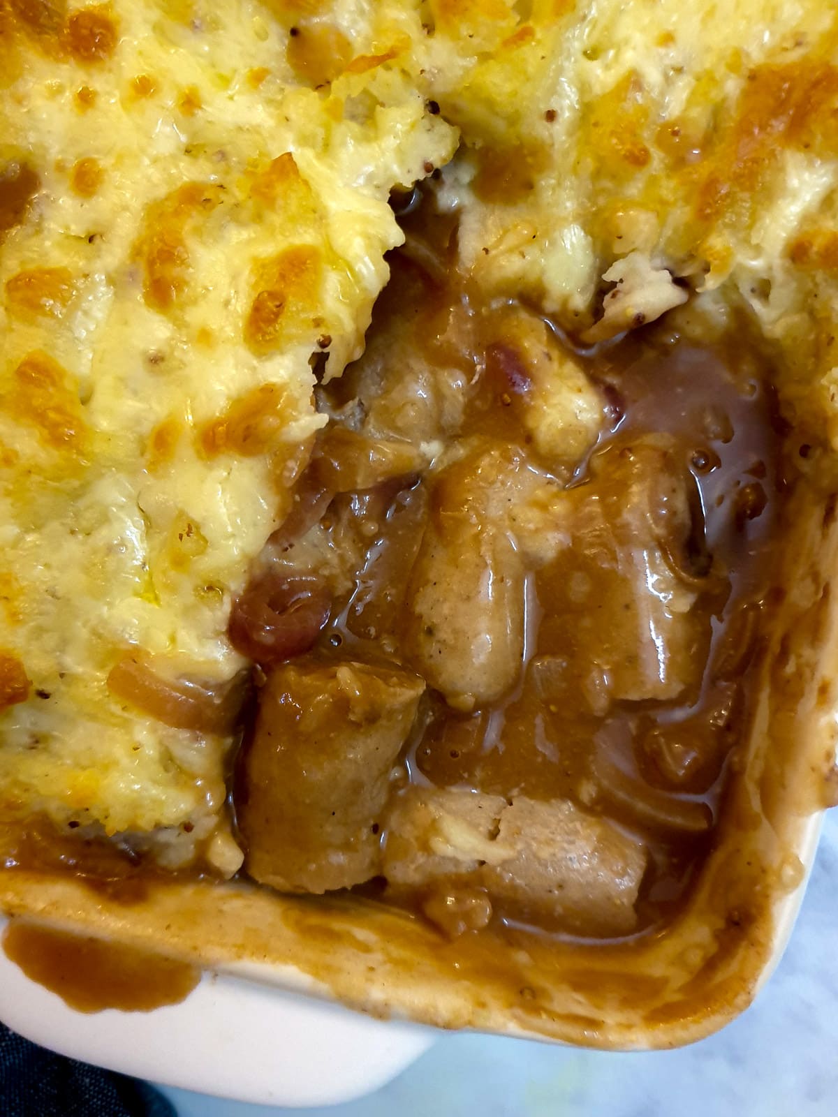 Pieces of sausages with onion gravy in the corner of a casserole dish, surrounded with cheesy mashed potatoes.