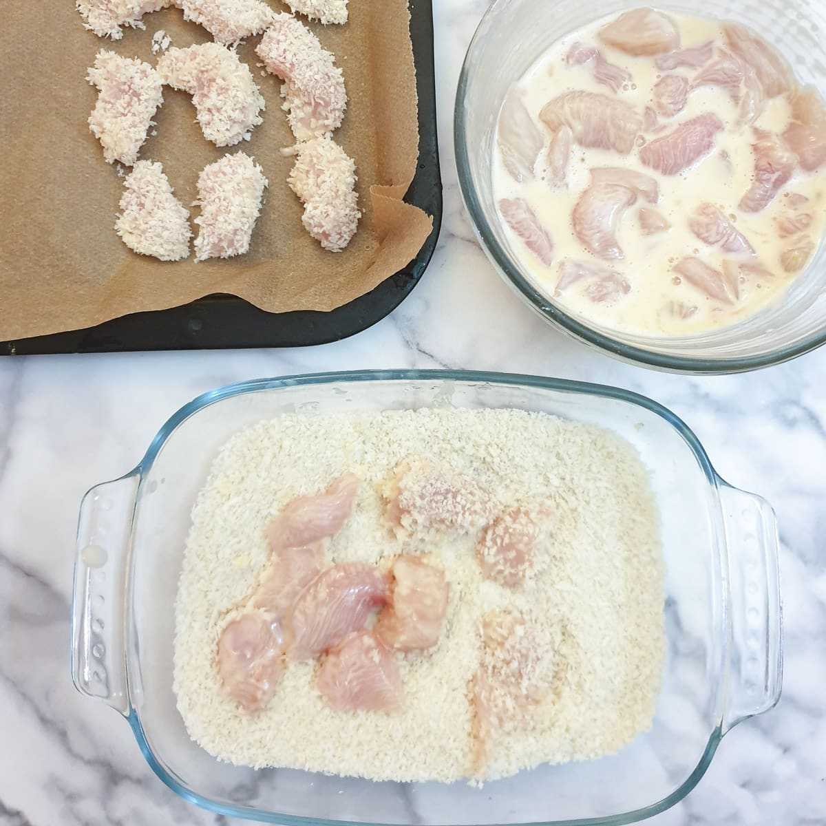 Pieces of chicken being coated in breadcrumbs and arranged on a baking tray.