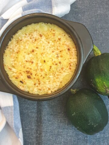A dish of baked gem squash on a blue tablecloth next to 2 whole raw gem squash.