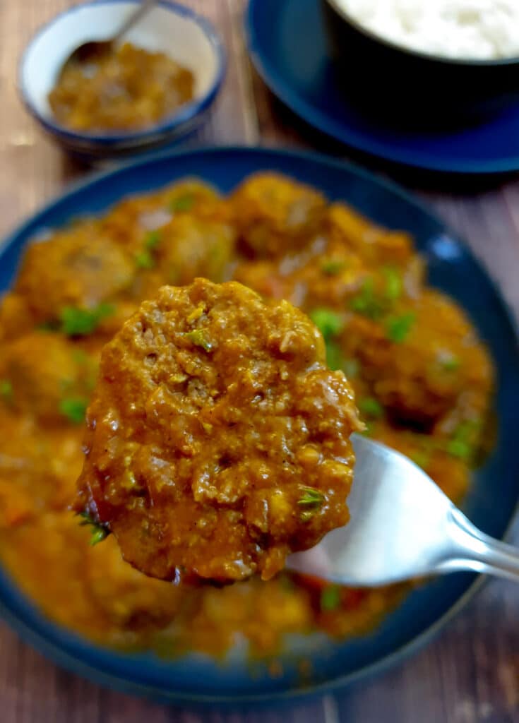 Close up of half a meatball on a fork held above the serving dish of curried meatballs.