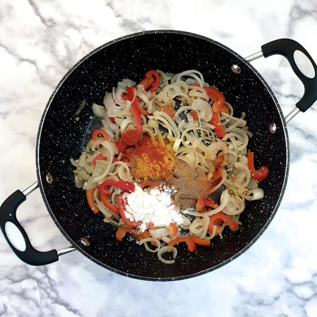 Spices and flour spooned onto cooked onions and red peppers in a frying pan.