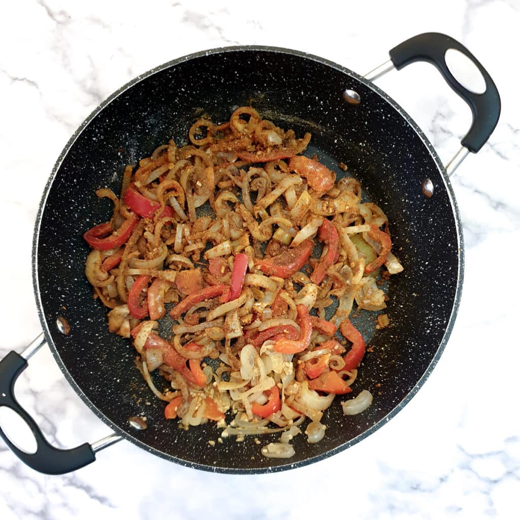 Spices and flour mixed with onions and red peppers in a frying pan.