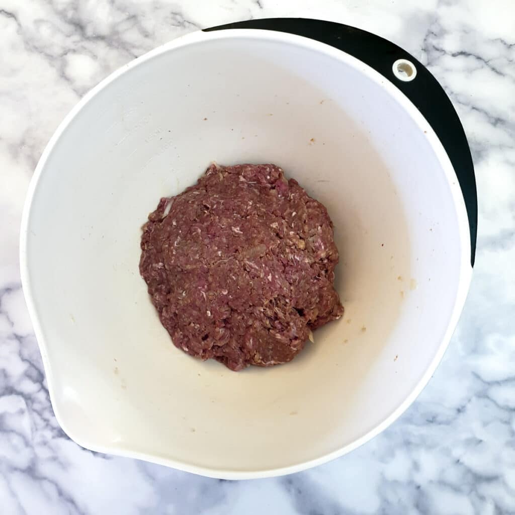 A white mixing bowl containing minced beef mixed with egg, spices and breadcrumbs, ready to be formed into meatballs.