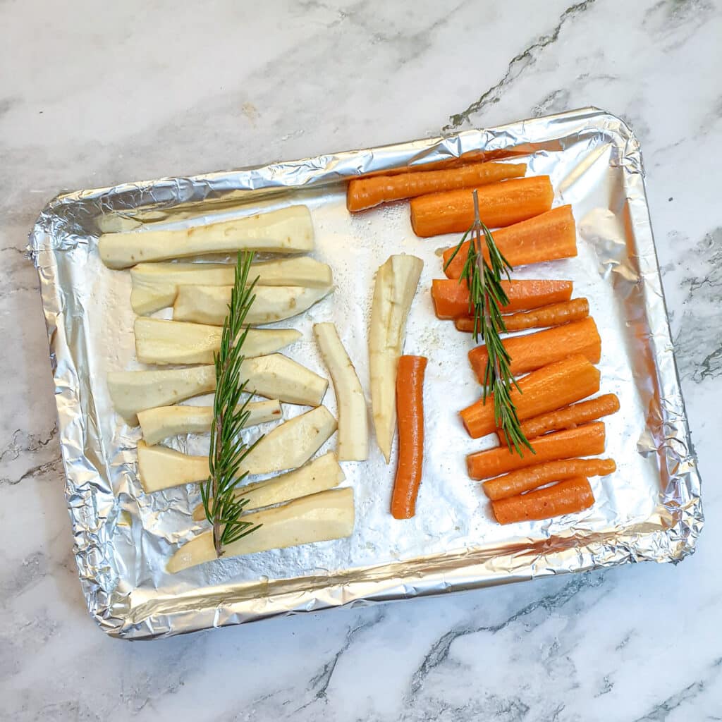 Carrots and parsnips arraned on a foil-lined baking tray, topped with rosemary.