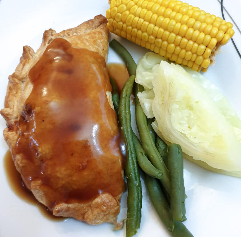 A Cornish pasty on a white plate, covered with gravy, with corn, beans and cabbage on the side.